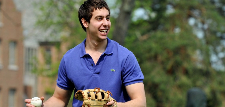 A white male student with brown hair wearing a blue collared shirt holding a baseball glove getting read to throw the baseball on the quad.